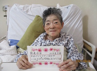 入居者が居室のベッドで記念撮影。笑顔で手書きメッセージボードを持つ。