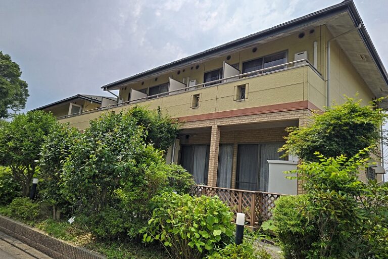 緑に囲まれた2階建ての介護施設の建物外観。バルコニー付きの現代的な施設。